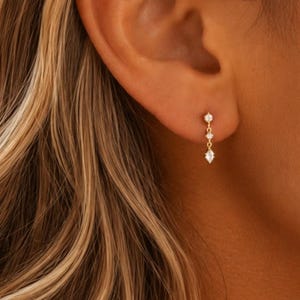 May include: A delicate gold drop earring featuring two small round clear stones set vertically, ending in a single marquise-shaped clear stone at the base. The earring is attached to a thin gold chain link, creating a subtle, dangling vertical design.