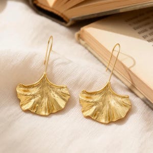 Puede incluir: Un par de pendientes dorados con forma de hojas de ginkgo. Los pendientes están sobre una superficie blanca, con un libro abierto en el fondo. Los pendientes tienen una superficie texturizada y cierre de gancho.