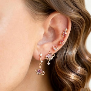 May include: A collection of gold-toned earrings featuring celestial and heart designs. The earrings include a dangling planet-shaped earring with a pink gemstone, heart-shaped studs, and a star-accented earring. The earrings are worn on an ear.