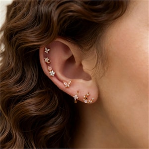 Puede incluir: Primer plano de una oreja adornada con múltiples pendientes dorados. Los pendientes presentan varios diseños, incluyendo flores, mariposas y una luna creciente, algunos con pequeñas gemas brillantes. La oreja se encuentra sobre un fondo de cabello castaño ondulado.
