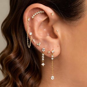 May include: An ear adorned with multiple gold earrings featuring clear gemstones. The earrings include studs, chain dangles, and a curved bar. The jewelry is set against the backdrop of wavy brown hair.