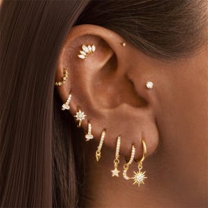 Può includere: Primo piano di un orecchio adornato da molteplici orecchini d'oro. Gli orecchini includono piccoli borchie, cerchi con ciondoli pendenti e un gruppo di pietre. I gioielli sono incastonati con pietre preziose trasparenti, creando un effetto scintillante.