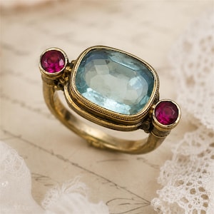 May include: A gold-toned ring featuring a large, rectangular, faceted light blue stone set in the centre, flanked by two smaller, round, faceted deep pink stones on either side. The metal band has a textured, aged finish with intricate detailing.