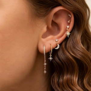 Puede incluir: Una colección de pendientes dorados con diseños celestiales. Los pendientes incluyen una estrella colgante, una media luna y un diseño de estrella, todos adornados con pequeñas gemas brillantes. Las perforaciones en la oreja también incluyen un pequeño perno y un rayo.