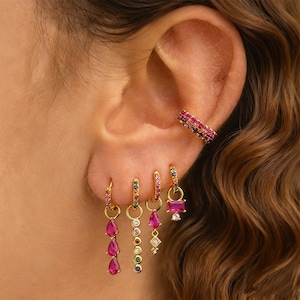 Puede incluir: Pendientes y puño de oreja dorados con piedras preciosas de colores. Los pendientes incluyen formas de lágrima, redondas y rectangulares con piedras rosas, moradas, verdes y azules. El puño de oreja está adornado con gemas rosas.