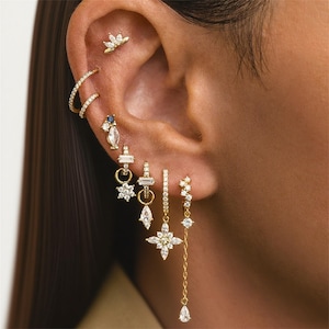 Peut inclure: Une oreille ornée de multiples boucles d'oreilles dorées. Les boucles d'oreilles comprennent de petits anneaux, des clous et des modèles pendants avec des pierres précieuses claires et bleues. Un petit clou de couronne en or est visible.