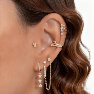 May include: A collection of gold-toned earrings and ear cuffs. The earrings include a small stud, a hoop with dangling stars, and a chain design. The ear cuffs feature a layered design with clear gemstones.
