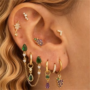 Pode incluir: Uma variedade de brincos dourados adorna uma orelha. Os brincos apresentam vários designs, incluindo uma estrela, um aglomerado de pedras preciosas e formas de gota. Alguns brincos têm pedras verdes e roxas, e alguns têm detalhes de corrente.