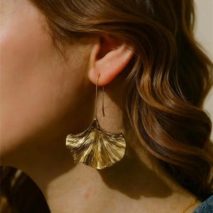 Puede incluir: Pendientes dorados con forma de hoja. Los pendientes tienen un gancho largo y delgado y una superficie detallada y texturizada. El diseño de la hoja recuerda a una hoja de ginkgo. Los pendientes son una pieza llamativa.