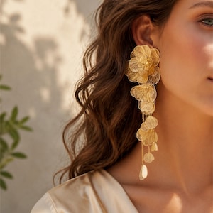 Könnte beinhalten: Goldfarbene, blütenförmige Ohrringe mit einem Kaskaden-Design. Die Ohrringe weisen mehrere Schichten aus zarten, blütenblattartigen Formen auf, die einen fließenden, eleganten Look erzeugen. Getragen von einer Person mit langen, welligen braunen Haaren und einem cremefarbenen Oberteil.