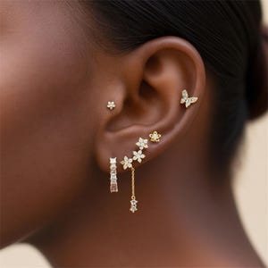 May include: A collection of gold earrings, including a small flower stud, a butterfly stud, and a series of floral and geometric designs. The earrings are adorned with small, sparkling gemstones, creating a delicate and elegant aesthetic.