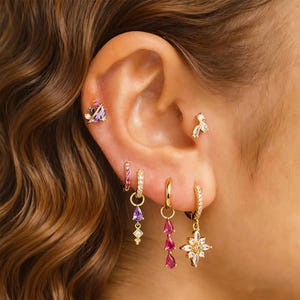 Può includere: Primo piano di un orecchio adornato da diversi orecchini d'oro. Gli orecchini presentano vari design, tra cui piccoli cerchi con gemme rosa, gemme a goccia viola e rosa pendenti e un orecchino a forma di fiore con gemme trasparenti.