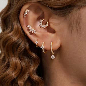 Könnte beinhalten: Nahaufnahme eines Ohrs, das mit mehreren goldenen Ohrringen geschmückt ist. Die Ohrringe weisen klare, funkelnde Steine in verschiedenen Designs auf, darunter Blumen- und geometrische Formen. Das Ohr ist vor einem Hintergrund aus welligem, hellbraunem Haar platziert.