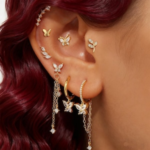 Könnte beinhalten: Ein Ohr ist mit einer Sammlung goldfarbener Ohrringe geschmückt, darunter Schmetterlings- und Blattmotive. Die Ohrringe sind mit klaren, funkelnden Akzenten verziert. Das Set umfasst Ohrstecker, Creolen und Hängeohrringe, die einen vielschichtigen und eleganten Look erzeugen.