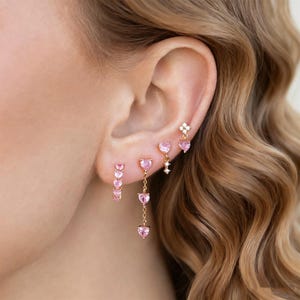 Puede incluir: Una colección de pendientes dorados con gemas rosas en forma de corazón. Los pendientes incluyen tachuelas, estilos colgantes y un diseño de racimo, mostrando una estética romántica y femenina. Los pendientes se muestran en una oreja.