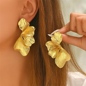 Puede incluir: Pendientes dorados con diseño floral. Los pendientes tienen un diseño de pétalos texturizados y en capas. Los pendientes están siendo sostenidos por una mano. Un accesorio llamativo.