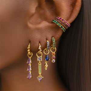 May include: An assortment of gold earrings and ear cuffs. The earrings showcase diverse gemstone designs, such as teardrops, squares, and triangles, in shades of purple, blue, green, and pink. The ear cuffs are embellished with rows of colourful gemstones.