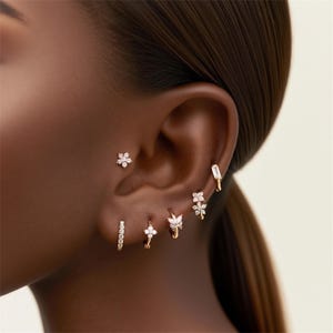 May include: A collection of gold earrings adorns an ear, featuring a variety of designs. These include a small flower stud, a rectangular bar, a small hoop with a row of gems, and several small hoops with floral and butterfly accents.