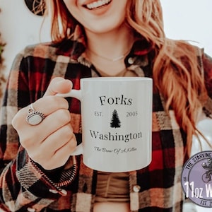 Może przedstawiać: Biały ceramiczny kubek z napisem "Forks Washington" i grafiką drzewa. Kubek zawiera również tekst "EST. 2005" i "The Brew Of A Killer". Kubek jest trzymany przez osobę noszącą koszulę w kratę.