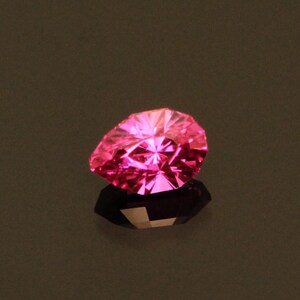 Puede incluir: Una gema rosa brillante, facetada en forma de pera. La gema es muy reflectante, con múltiples facetas que capturan la luz. Se asienta sobre una superficie oscura y reflectante, creando un efecto de espejo. El fondo es de un tono neutro y apagado.