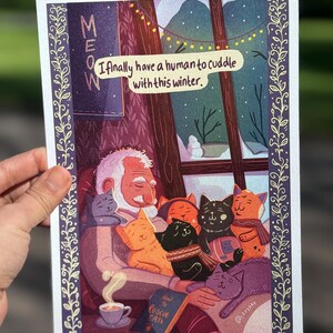 May include: A print featuring a cozy winter scene with an elderly person surrounded by several cats. The text reads "I finally have a human to cuddle with this winter." The word "MEOW" is also visible. A book titled "How to Rescue Cats" is in the image.