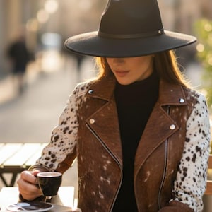 Könnte beinhalten: Ein schwarzer Fedora-Hut mit breiter Krempe. Eine braune Lederjacke mit weiß-braunen gefleckten Ärmeln. Die Person trägt einen schwarzen Rollkragenpullover und hält eine Tasse Kaffee. Die Umgebung ist im Freien.