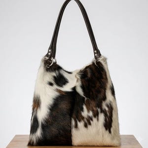 May include: A hobo-style shoulder bag featuring a cowhide pattern with patches of white, black, and dark brown fur. The bag includes two dark brown leather shoulder straps attached with silver-toned metal rings on each side of the top opening.