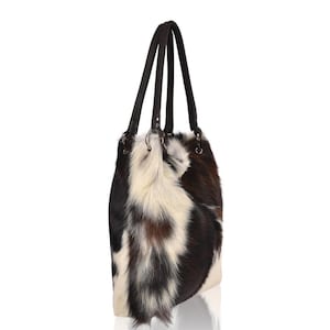 May include: A tote bag featuring a multi-colored cowhide exterior with patches of white, dark brown, and black fur. The bag includes two dark brown leather shoulder straps attached with silver-toned metal rings on the top sides of the bag.