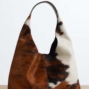 May include: A large, brown, white, and black cowhide shoulder bag with a black leather handle. The bag has a soft, slouchy shape and a natural, textured appearance. The bag is made of genuine cowhide.