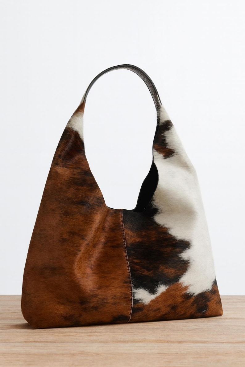 May include: A large, brown, white, and black cowhide shoulder bag with a black leather handle. The bag has a soft, slouchy shape and a natural, textured appearance. The bag is made of genuine cowhide.