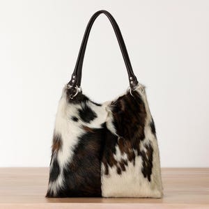 May include: A hobo-style shoulder bag crafted from cowhide featuring a natural tri-color pattern of white, brown, and black patches. The bag includes two dark brown leather handles attached with silver-toned metal rings and rivets.