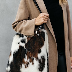 May include: A shoulder bag featuring a cowhide pattern with patches of white, black, and brown fur. The bag has a black leather strap attached to a silver-toned metal ring hardware. The texture of the bag is visibly hairy and soft to the touch.