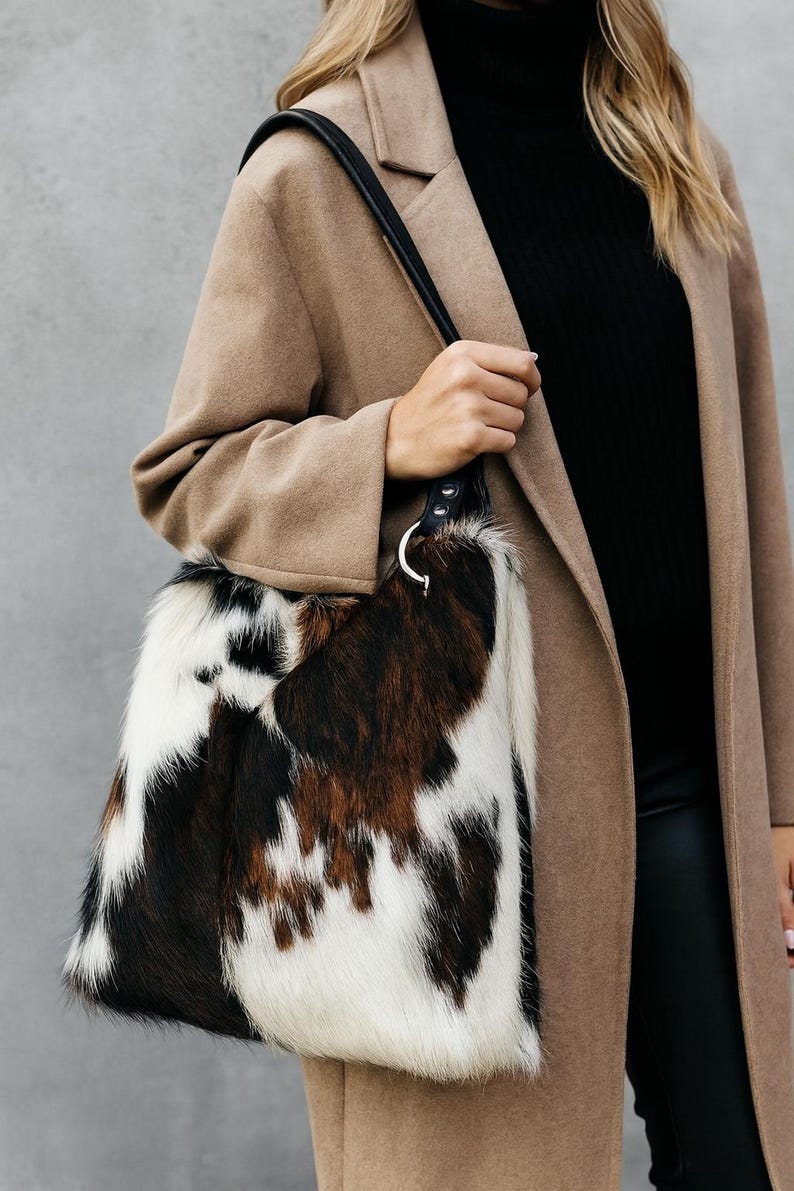 May include: A shoulder bag featuring a cowhide pattern with patches of white, black, and brown fur. The bag has a black leather strap attached to a silver-toned metal ring hardware. The texture of the bag is visibly hairy and soft to the touch.