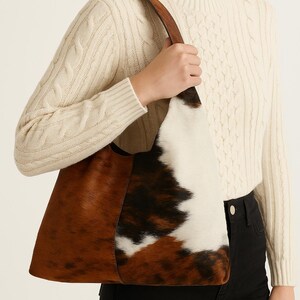 May include: A cowhide tote bag in shades of brown, black, and white, with a leather strap. The bag is held by a person wearing a cream cable knit sweater and black trousers. The bag's design features a mix of brown, black, and white fur.