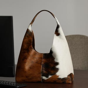 May include: A large cowhide shoulder bag with a curved handle. The bag has a patchwork design with brown, white, and black patches. The bag is on a wooden desk.