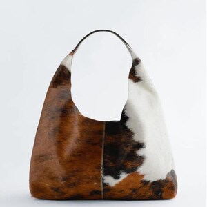 May include: A large shoulder bag made from cowhide, featuring a curved handle. The bag has a patchwork design with brown, white, and black fur. The bag is a neutral colour and has a simple, modern design.