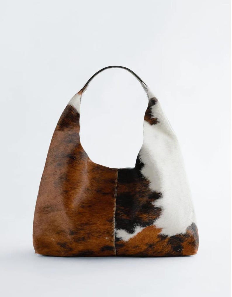 May include: A large shoulder bag made from cowhide, featuring a curved handle. The bag has a patchwork design with brown, white, and black fur. The bag is a neutral colour and has a simple, modern design.