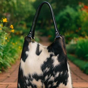 May include: A hobo-style handbag featuring a black and white cowhide pattern with a textured fur finish. The bag includes two black leather shoulder straps attached with gold-toned metal rings and rivets, providing a structured yet soft silhouette.