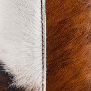 May include: Close-up of a cowhide rug with a stitched seam. The rug features a white and brown colour scheme. The white section has long, soft fur, while the brown section has shorter, textured fur.