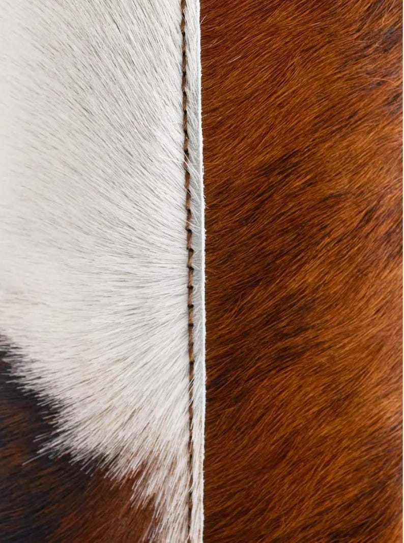 May include: Close-up of a cowhide rug with a stitched seam. The rug features a white and brown colour scheme. The white section has long, soft fur, while the brown section has shorter, textured fur.