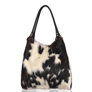 May include: A hobo-style shoulder bag featuring a cowhide pattern with irregular black and white patches. The bag has two dark brown leather handles attached to the top with circular metal rings, providing a structured yet soft silhouette.