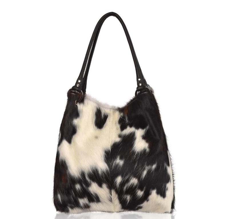 May include: A hobo-style shoulder bag featuring a cowhide pattern with irregular black and white patches. The bag has two dark brown leather handles attached to the top with circular metal rings, providing a structured yet soft silhouette.