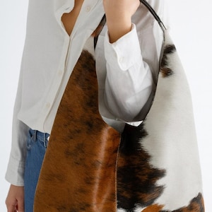 May include: A large shoulder bag with a cowhide pattern. The bag features a mix of brown, white, and black patches. The strap is black, and the bag is held by a person wearing a white shirt and blue jeans.