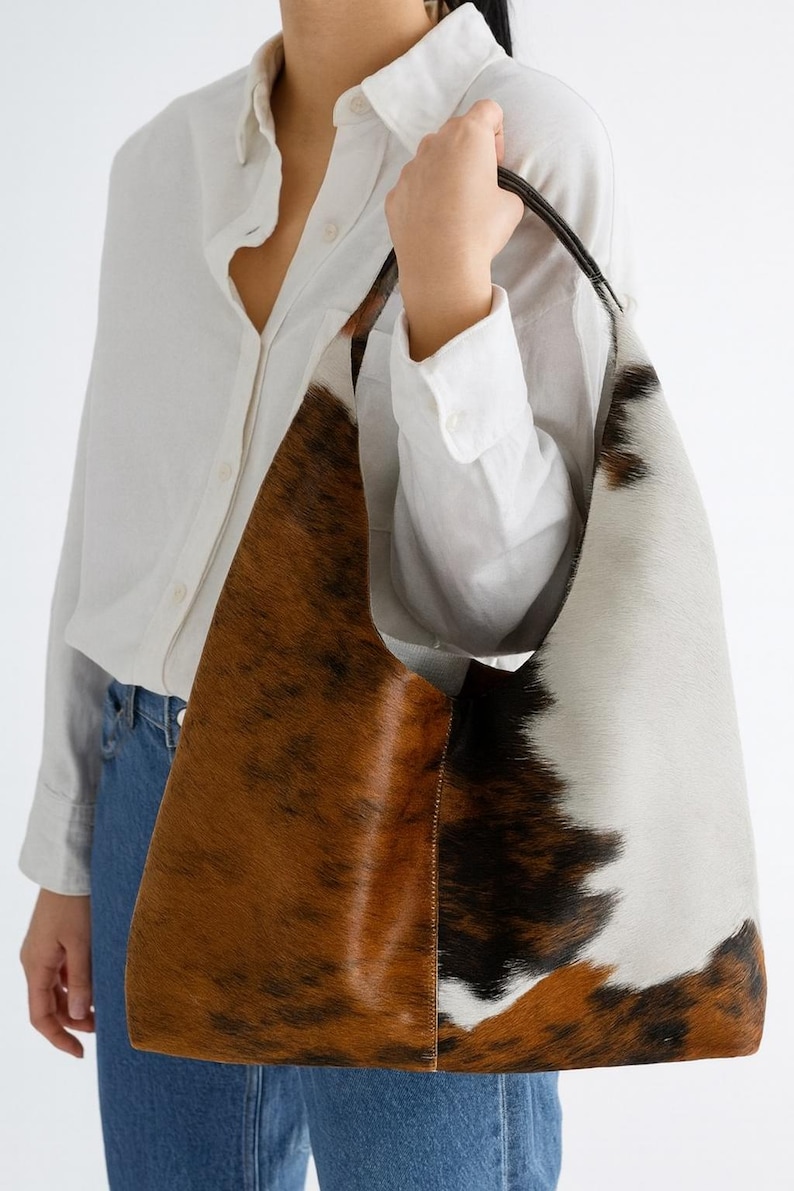 May include: A large shoulder bag with a cowhide pattern. The bag features a mix of brown, white, and black patches. The strap is black, and the bag is held by a person wearing a white shirt and blue jeans.
