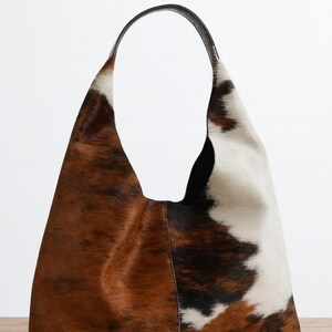 May include: A large, brown, white, and black cowhide shoulder bag with a black leather handle. The bag has a soft, slouchy shape and a natural, textured appearance. The bag is made of genuine cowhide.