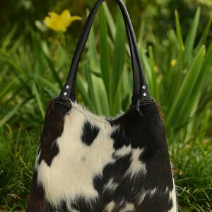 May include: A hobo-style handbag featuring a cowhide exterior with a black, white, and brown spotted pattern. The bag includes two black leather shoulder straps attached with silver-toned metal hardware and circular rings at the top corners.