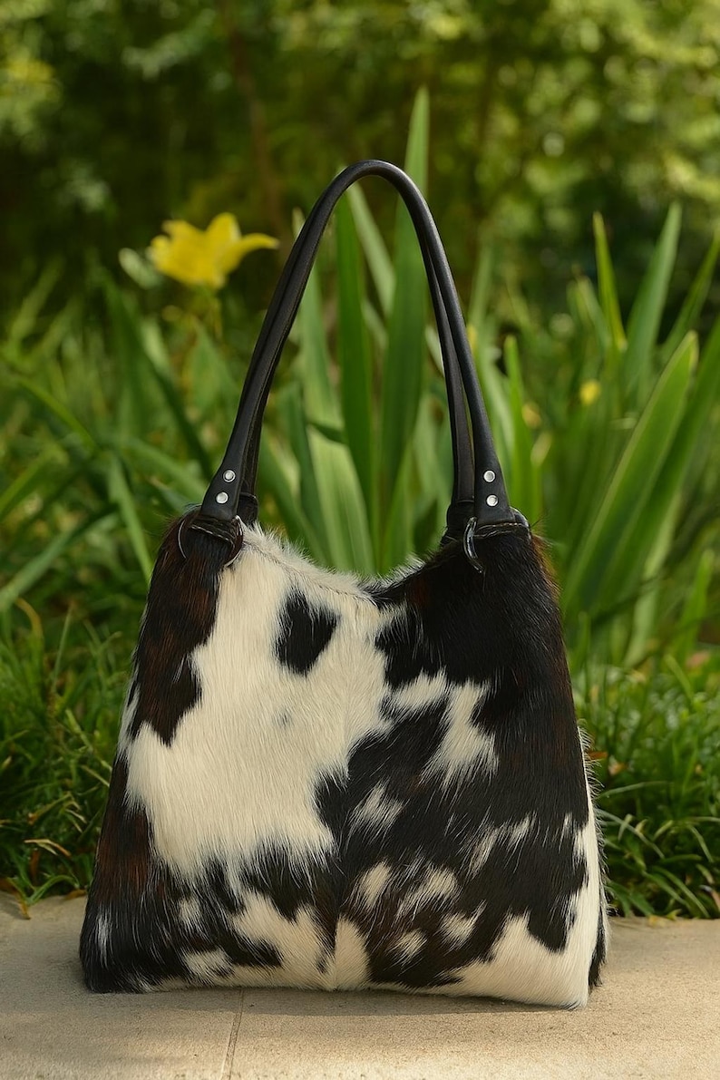 May include: A hobo-style handbag featuring a cowhide exterior with a black, white, and brown spotted pattern. The bag includes two black leather shoulder straps attached with silver-toned metal hardware and circular rings at the top corners.