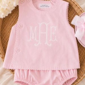 May include: A light pink sleeveless top and matching bloomer set. The top features a scalloped edge, two buttons on the shoulder, two buttons on the side, and a white embroidered monogram. A label reads Palmetto Stitch Company.