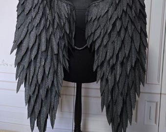 Könnte beinhalten: Große, dunkelgraue Engelsflügel mit einem gefiederten Design. Die Flügel werden auf einer schwarzen Schaufensterpuppe ausgestellt, um ihre Größe und Details zu zeigen. Die Flügel bestehen aus einem strukturierten Material, und die Federn haben spitze Spitzen.