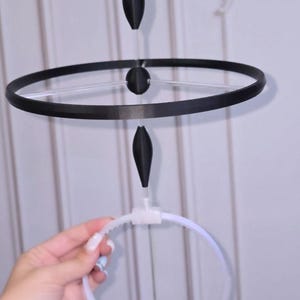 May include: A black halo headband featuring a thin, circular black ring suspended horizontally above a white plastic headband. The structure includes two black, pointed, spindle-shaped ornaments positioned vertically on a clear central rod.